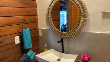 Un baño con pared de madera, un espejo redondo con marco de bambú, un lavamanos blanco, un perchero con una toalla azul y un pequeño jarrón con una flor rosa.