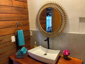 Ein Badezimmer mit einer Holzwand, einem runden Spiegel mit Bambusrahmen, einem weißen Waschbecken, einem Handtuchhalter mit blauem Handtuch und einer kleinen Vase mit einer rosa Blume.