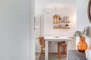 Een moderne keuken met witte kastjes, een wit aanrecht en twee rieten stoelen. Boven het aanrecht zijn planken met decoratieve objecten en een potplant.