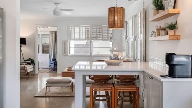 A modern kitchen with a central island, wooden stools, a hanging light fixture, and built-in shelving with plants. - House of Rio, Seaside Getaway (Jensen Beach)