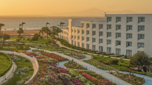 Un hotel con diseño moderno, que cuenta con un jardín ajardinado con diversas plantas y flores, un camino que lleva a la playa, y vistas al océano y a las montañas en la distancia.