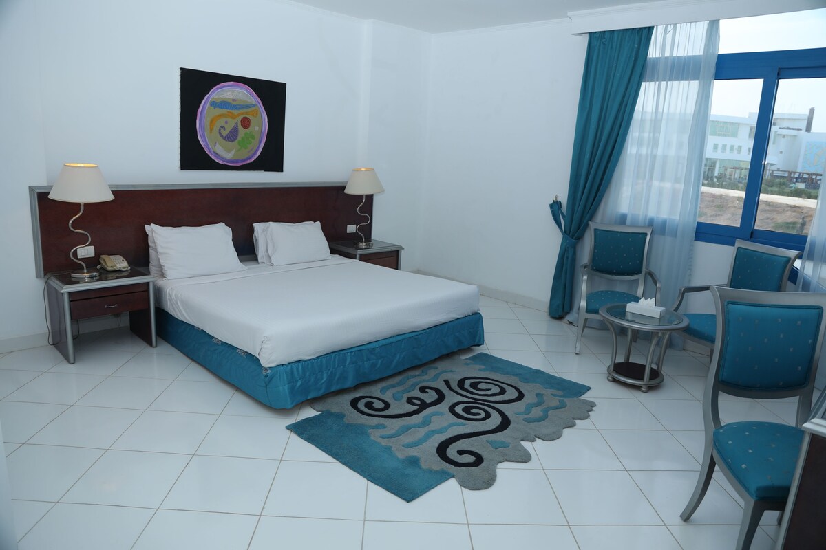Habitación de hotel con cama, dos mesitas de noche con lámparas, una mesita redonda pequeña con una silla y vistas al exterior a través de una ventana con cortinas azules.