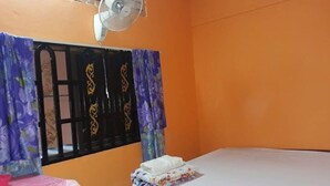 A room with orange walls, a bed with a white mattress and a red headboard, a small table with a pink tablecloth, a fan on the ceiling, and a window with purple curtains.