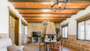 A cozy living room with a stone fireplace, wooden ceiling beams, and a dining area with a table set for four. - Nice home in Casariche (Casariche)