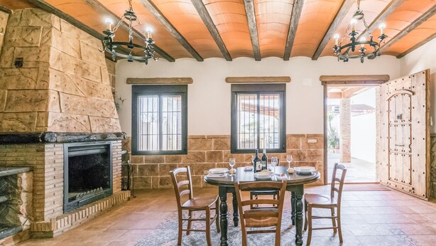 A dining area with a stone fireplace, wooden ceiling beams, and a round table set for four. - Nice home in Casariche (Casariche)