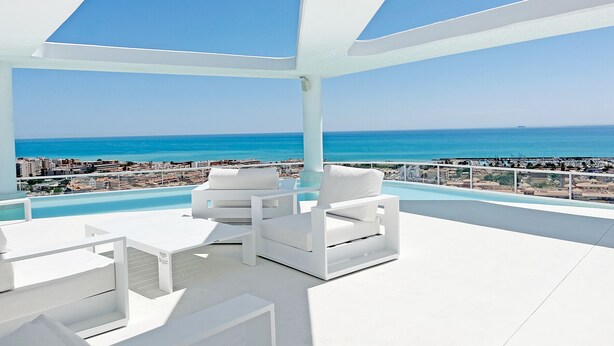 Un moderno espacio lounge al aire libre con mobiliario blanco, piscina y vistas al mar y al paisaje urbano.