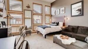 Ein Schlafzimmer mit einem Bett, einer Couch, einem Schreibtisch und einem Stuhl. Es gibt mehrere Fenster mit Jalousien, eine Lampe und gerahmte Bilder an der Wand.
