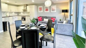 Una cocina moderna con armarios blancos, un comedor con mesa para cuatro, y un sofá gris en la sala.