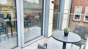 Un balcón con puerta de cristal, una mesa redonda con planta en maceta y asientos para exteriores.