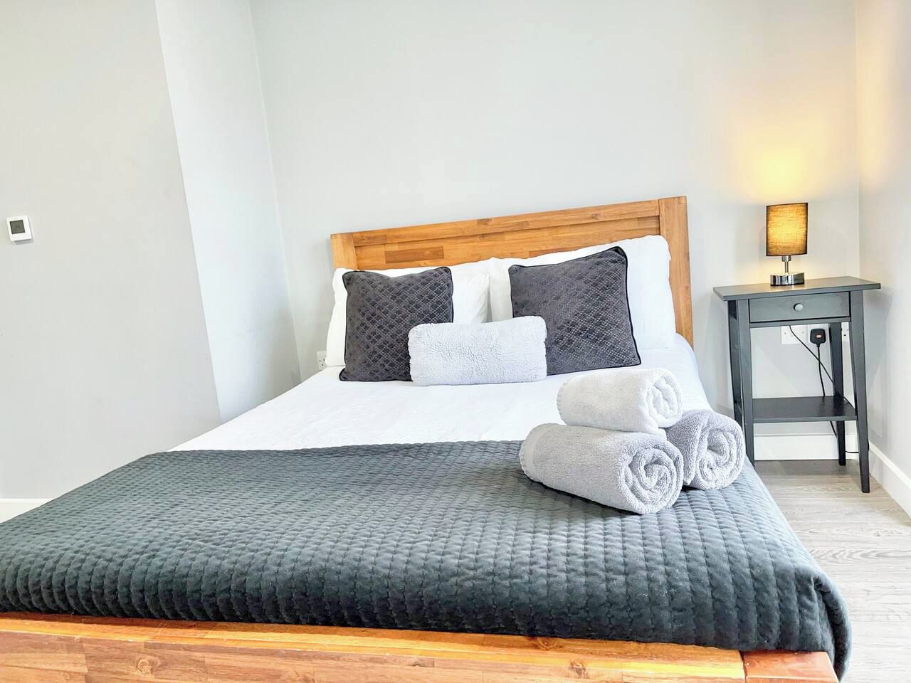 A neatly made bed with a grey quilt, two pillows, and rolled towels.