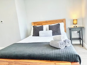 A neatly made bed with a grey quilt, two pillows, and rolled towels.