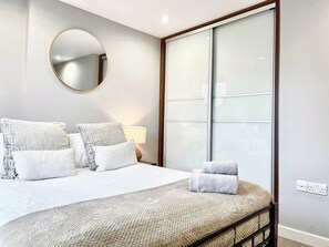 A hotel room with a bed, pillows, a round mirror, a bedside lamp, and a large window with frosted glass.