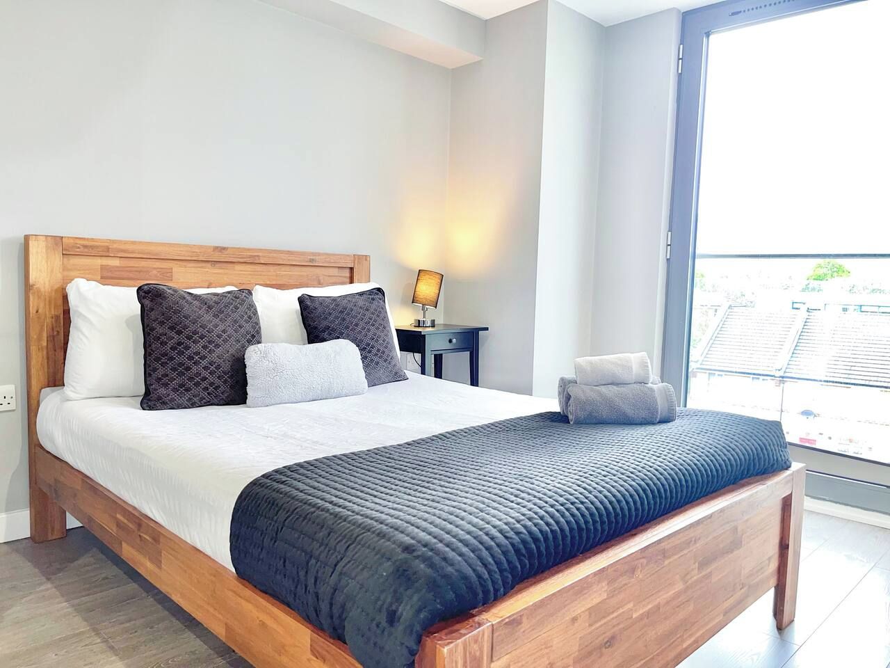 A modern bedroom with a wooden bed, white bedding, and a view of the city through a large window.