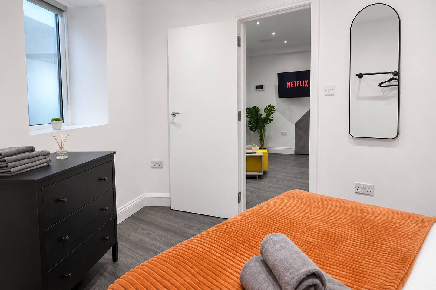 Ein modernes Schlafzimmer mit Bett, Kommode, Fenster und einer Tür zu einem anderen Raum.