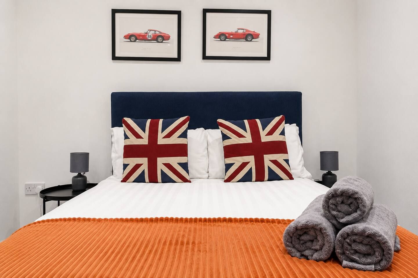 En seng med et hvidt og orange stribet sengetæppe, to puder med Union Jack-motiv, og to billeder i rammer på væggen over sengen.