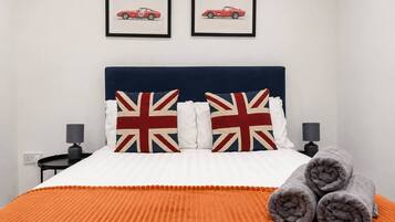 En seng med et hvidt og orange stribet sengetæppe, to puder med Union Jack-motiv, og to billeder i rammer på væggen over sengen.