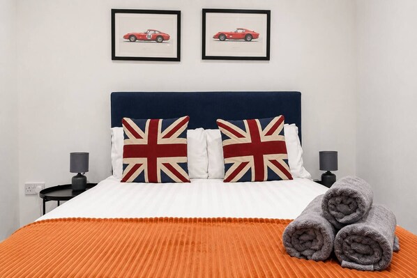 Una cama con colcha a rayas blanca y naranja, dos almohadas con diseño de la bandera del Reino Unido y dos cuadros enmarcados en la pared de arriba.