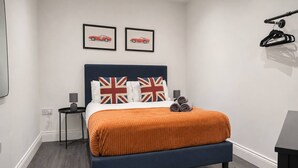 Un dormitorio con una cama, dos cuadros en la pared, una mesita de noche con una lámpara y un perchero negro en la pared.
