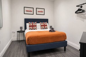 A bedroom with a bed, two framed pictures on the wall, a bedside table with a lamp, and a black hanger on the wall.