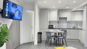 Een moderne woonkamer met een flatscreen-tv, een eethoek met tafel en stoelen, een keuken met witte kasten en apparatuur, en een afvalbak.
