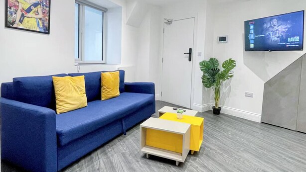 Una sala de estar moderna con un sofá azul, cojines amarillos, una pequeña mesa de centro, una planta en maceta y un televisor de pantalla plana montado en la pared.