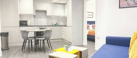 Una sala de estar moderna con un sofá azul, un comedor con sillas y una mesa, y una cocina con armarios blancos.