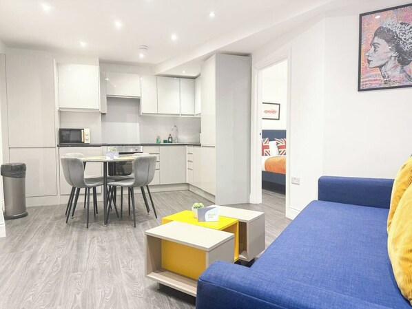 A modern living room with a blue sofa, a dining area with chairs and a table, and a kitchen with white cabinets.