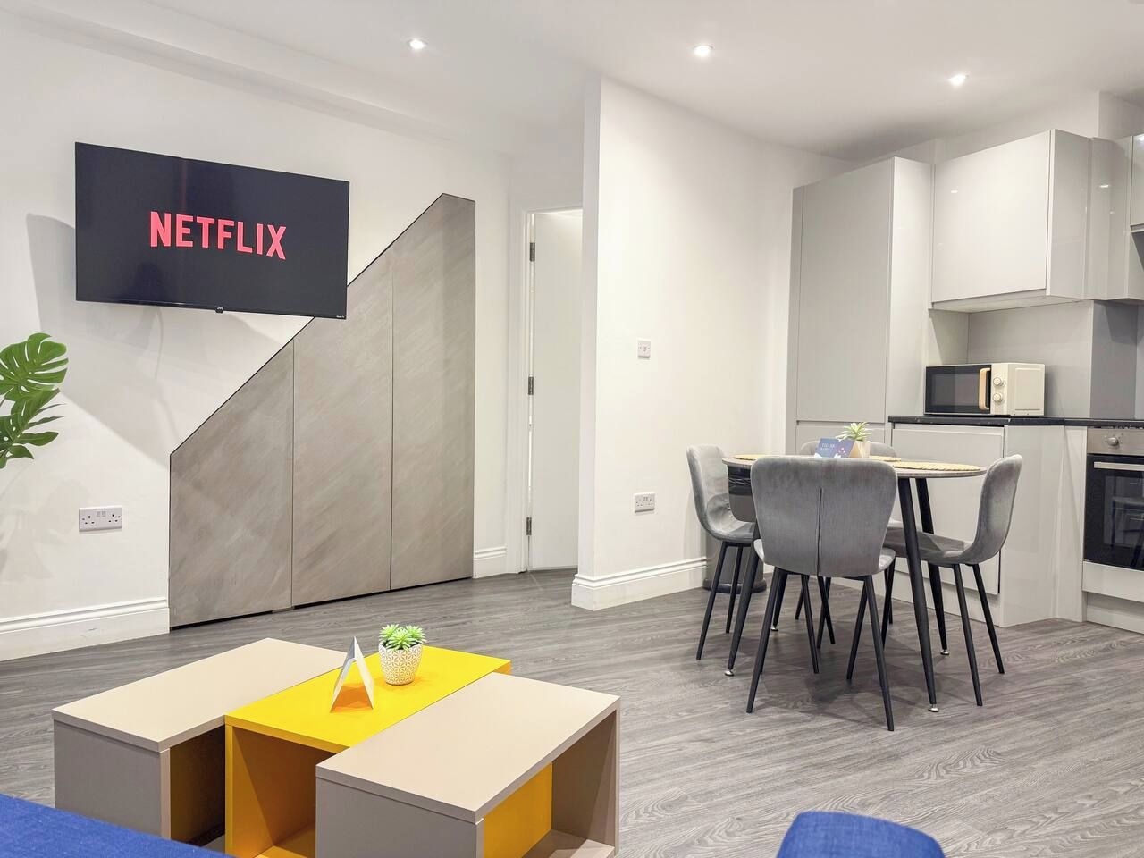 En moderne stue med et fladskærms-tv, der viser 'NETFLIX', et spiseområde med et lille bord og stole, og et køkken med skabe og en mikrobølgeovn.