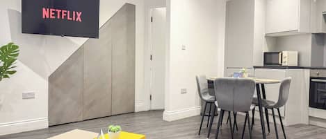 A modern living room with a flat-screen TV displaying 'NETFLIX,' a dining area with a small table and chairs, and a kitchen area with cabinets and a microwave.
