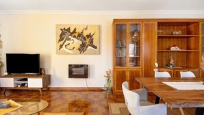 Una sala de estar moderna con un televisor de pantalla plana, un mueble de madera con puertas de vidrio, una mesa de comedor con sillas y un cuadro abstracto de gran tamaño en la pared.