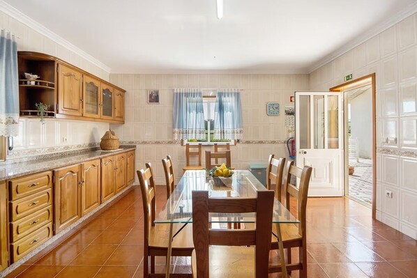 A spacious kitchen with wooden cabinets, a dining table with chairs, and a view of the garden through the window. - Villa da Floresta (Vila do Conde)