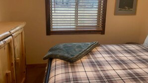 Ein Schlafzimmer mit kariertem Überwurf, einem Fenster mit Jalousien, einem gerahmten Bild an der Wand und einer Holzkommode.