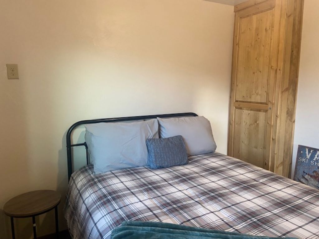 Een enkel bed met een geruit sprei en twee kussens, een klein rond tafeltje, en een houten deur in een kamer met effen muren.