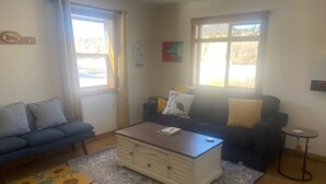 Ein Wohnzimmer mit einer Couch, einem Couchtisch und einem kleinen runden Tisch. Zwei Fenster sorgen für natürliches Licht.