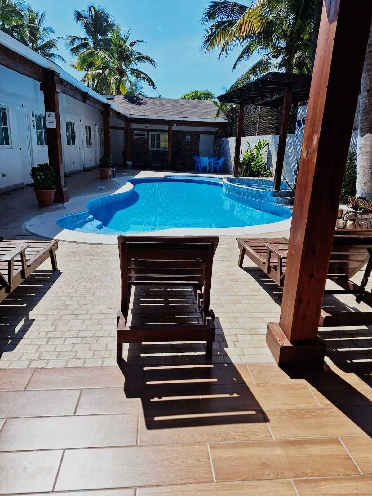 A swimming pool with a wooden deck and lounge chairs, surrounded by a covered patio area with palm trees.