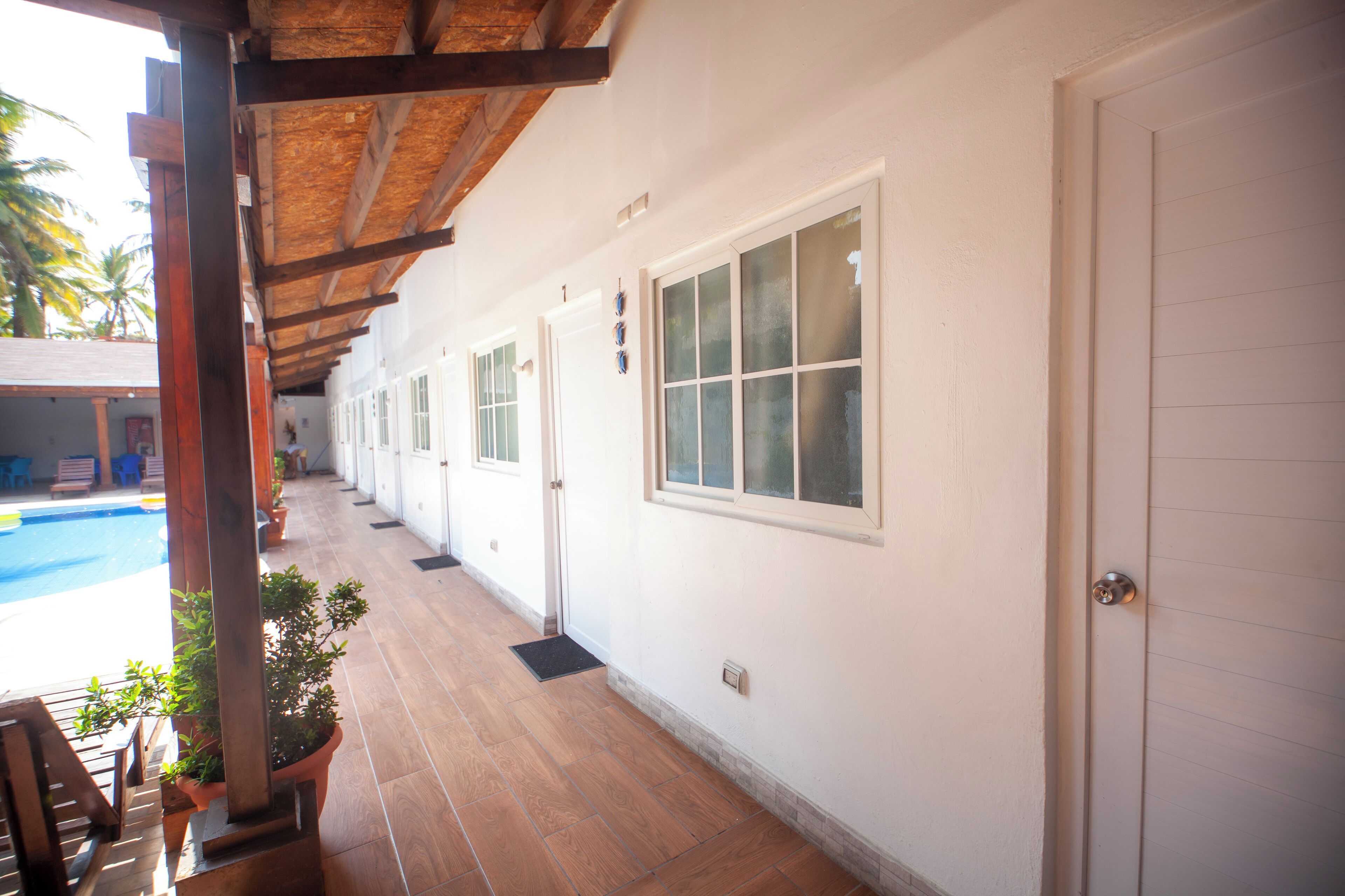 A covered corridor with doors and windows, potted plants, and a pool area visible in the background.