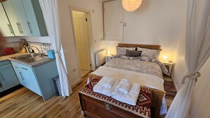 Een slaapkamer met een bed, nachtlampjes, een houten dressoir en een keukenhoek met een spoelbak en kastjes.
