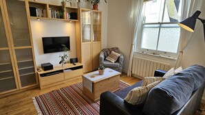 A modern living room with a flat-screen TV, wooden shelving, a grey armchair, and a patterned rug. - No 1 -  an apartment that sleeps 2 guests  in 1 bedroom (St Mawes)