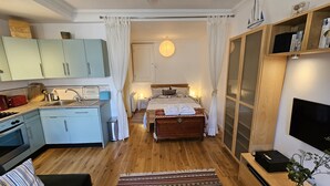 A compact hotel room with a kitchenette, a bed with a patterned bedspread, a bedside table with a lamp, and a large window with sheer curtains. - No 1 -  an apartment that sleeps 2 guests  in 1 bedroom (St Mawes)