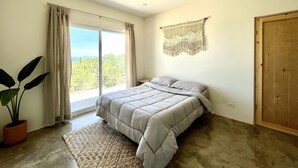 Een slaapkamer met een bed, een plafondventilator, een groot raam met gordijnen, een macramé wanddecoratie en een houten deur.