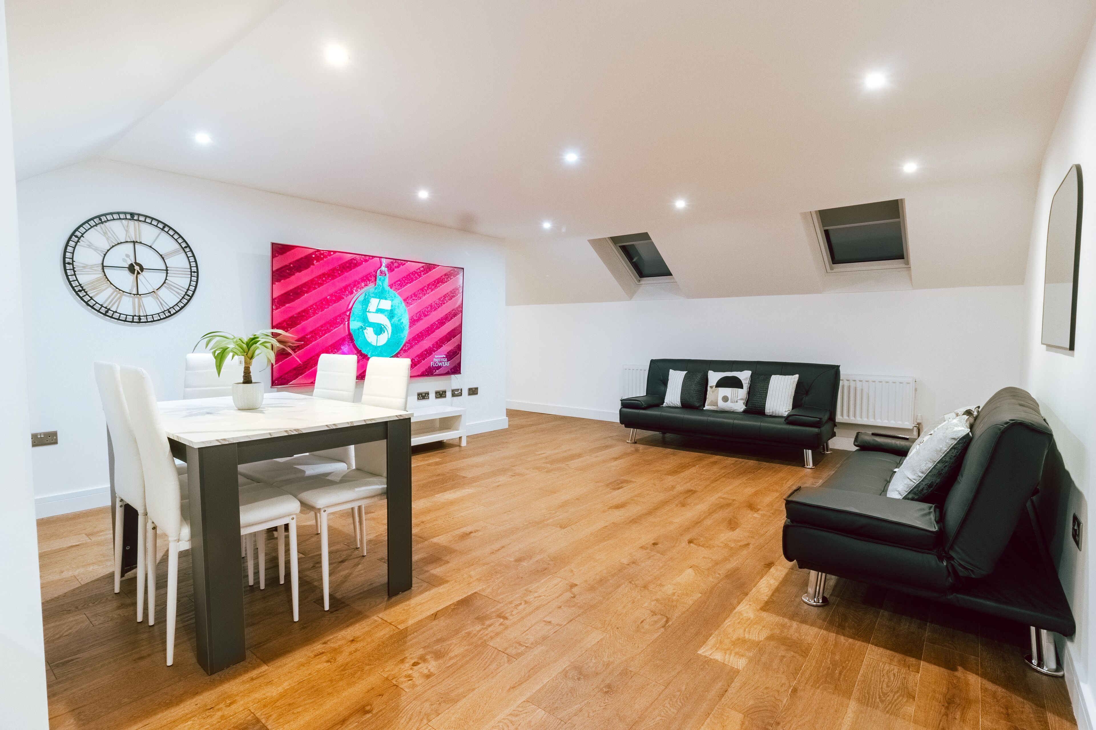 Un salon moderne comprenant un coin repas, un canapé noir, une grande horloge murale et un téléviseur fixé au mur.