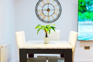 A dining area with a table, chairs, a potted plant, a wall clock, and a TV screen displaying a nature scene. - Remarkable, Penthouse - 3-bed Apartment in Purley (Purley)