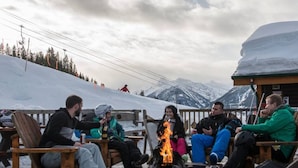 A group of people sitting around a campfire in a snowy outdoor setting with mountains in the background. - Moguls | Lofted Ski in Ski Out w/ Hot Tub Access (Panorama)