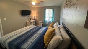 A bedroom with a bed, a ceiling fan, a TV mounted on the wall, a bedside table with a lamp, and a window with curtains. - Historic Peaceful Apt near VSU & SGMC (Valdosta)