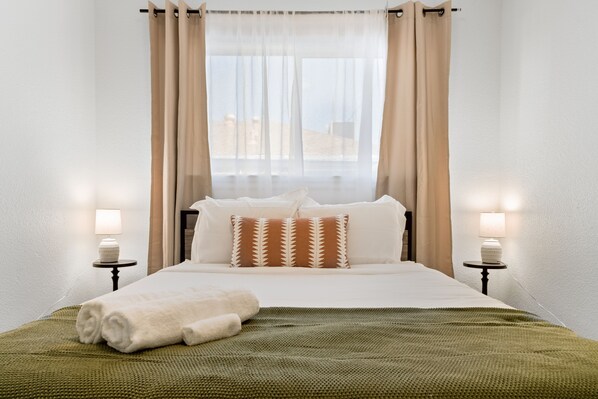 A neatly made bed with a green blanket and white towels, two bedside lamps, and a window with sheer curtains.