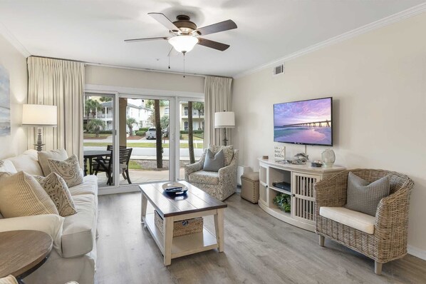 Un salón amplio con ventilador de techo, un televisor grande y vistas a una playa a través de puertas de cristal.
