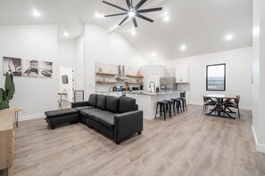Un salon moderne avec un canapé d’angle, un coin repas avec une table et des chaises, une cuisine avec des meubles blancs et un îlot central, et un ventilateur de plafond.