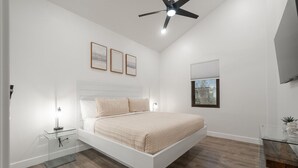 Une chambre à coucher moderne, avec un lit, des tables de chevet, un ventilateur de plafond et des œuvres d’art encadrées accrochées au mur.