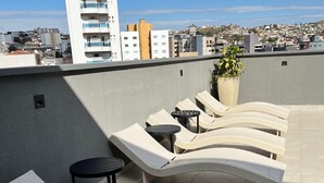 En takterrass med solstolar och ett litet bord, med utsikt över en stadssiluett med byggnader och en klar himmel.