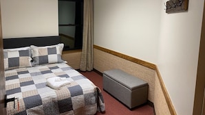 Una habitación de hotel con cama, ventana con cortinas, mesita de noche y un pequeño banco.